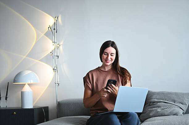 Woman messaging on smartphone while working on laptop, sitting comfortably on a sofa, lamp shades